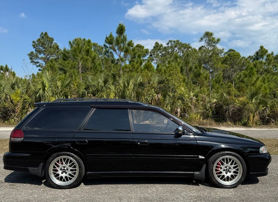 A 1996 Subaru Legacy Wagon GT-B in black, parked outdoors, showcasing its sleek, elongated body with sporty side skirts and factory alloy wheels.