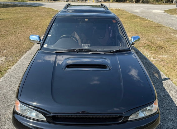 The front fascia of a 1996 Subaru Legacy Wagon GT-B, featuring its signature twin-turbo hood scoop, rectangular headlights, and a bold Subaru grille.