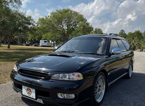 The front fascia of a 1996 Subaru Legacy Wagon GT-B, featuring its signature twin-turbo hood scoop, rectangular headlights, and a bold Subaru grille.