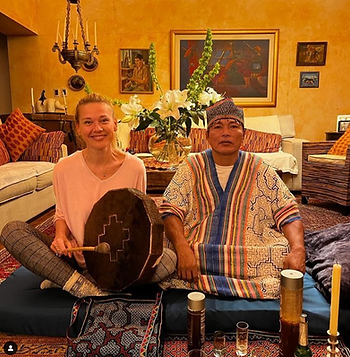 A blonde woman holding the ceremonial drum, sitting next to a peruvian shaman with the implements needed for an ayahuasca ceremony in front.