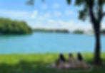 Three women seated on the grass by a serene lake, enjoying a peaceful moment in nature & just being