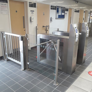 Two stainless-steel coin-operated tripod turnstiles at the entrance of a public facility, with payment instructions, guide rails, tiled flooring, and restroom doors visible in the background.