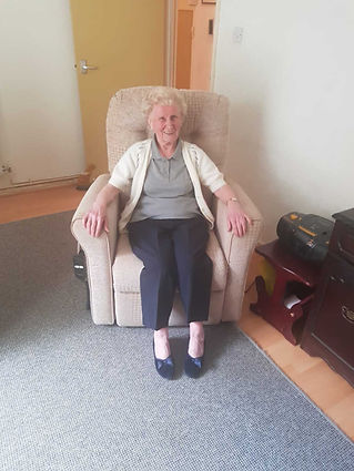 Elderly woman with short hair smiling, seated in a beige recliner in a cozy room