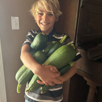 Young boy smiling holding many freshly picked, very large zucchini, harvest season. Long Valley Acres