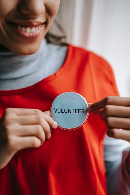 Smiling person holds a badge reading "VOLUNTEER." They wear a red shirt. Bright, positive mood with a blurred background.