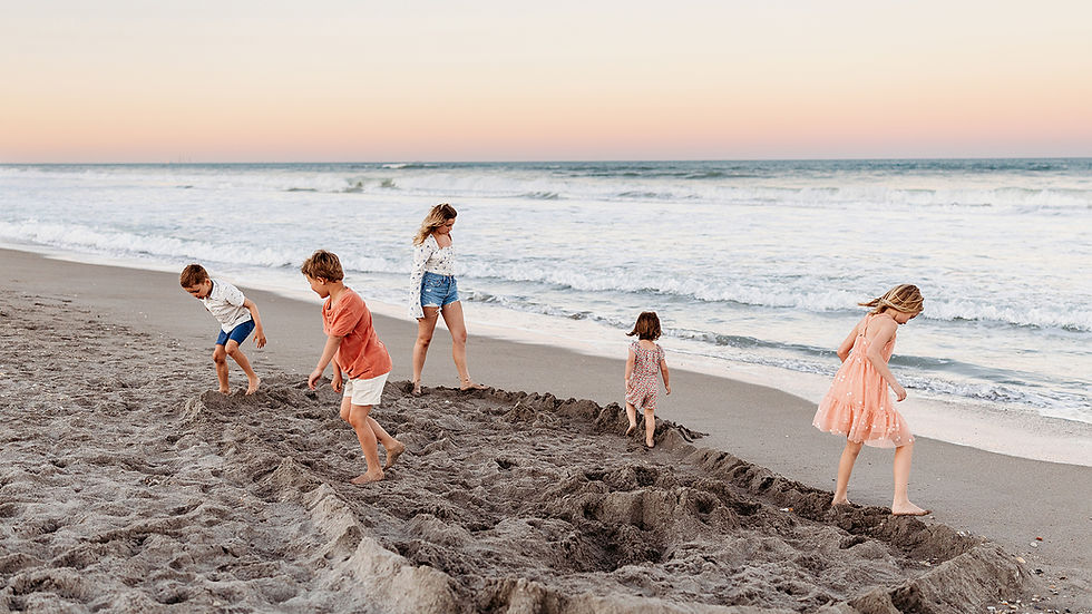 Children play in the sand at a beach during sunset. The sky is pink-orange and waves gently crash in the background.
