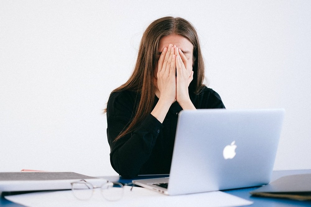 White woman with hands over her face in front of MacBook.