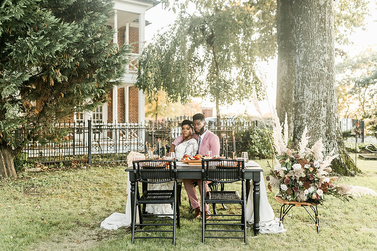A couple sits at a set table among lush trees in a picturesque setting.