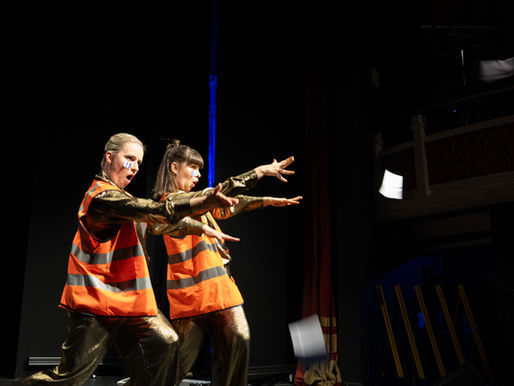 two people stand on stage in gold jumpsuits and orange high vis and throw paper into the audience
