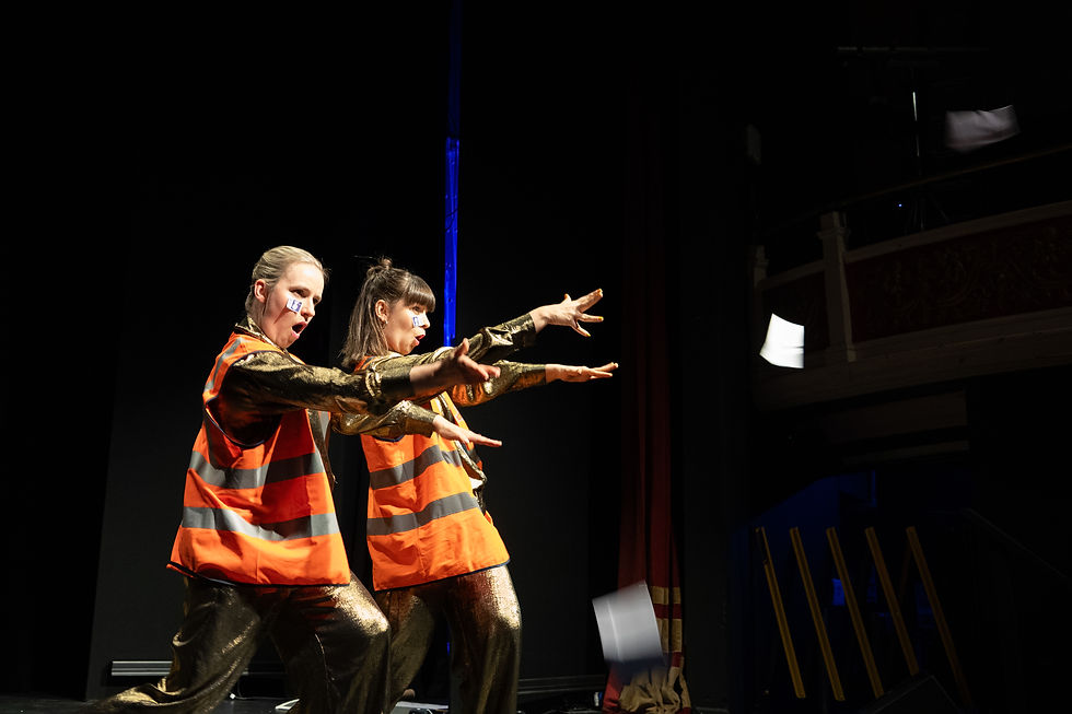 two people stand on stage in gold jumpsuits and orange high vis and throw paper into the audience