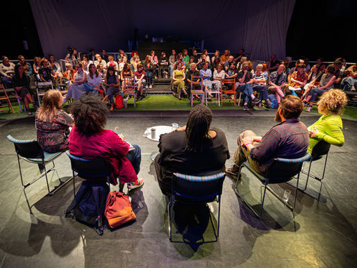 a panel of people sit on a stage talking, the camera behind them pointing at an audience listening to the pannelists