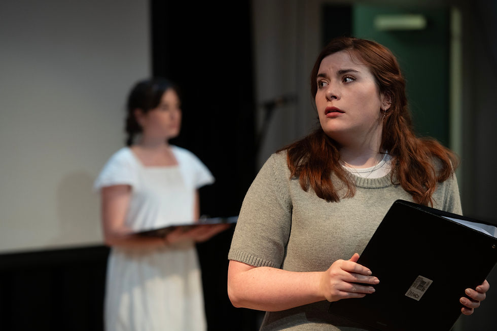 two people on stage. One stood close to the camera holding a script and looking scared of stage. Another standing at the back in a white blouse, blurred in the background