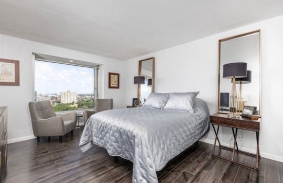 Elegant bedroom in chic condo with lounge chairs in front of window with gold vertical mirrors and tall, dark shade lamps atop rich wood night stands with dark luxury vinyl plank flooring.