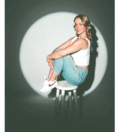 Eine junge Frau sitzt auf einem Hocker und hat die Füße ebenfalls auf dem Hocker abgestellt. Sie wird von einem Scheinwerfer angeleuchtet und lächelt in die Kamera.