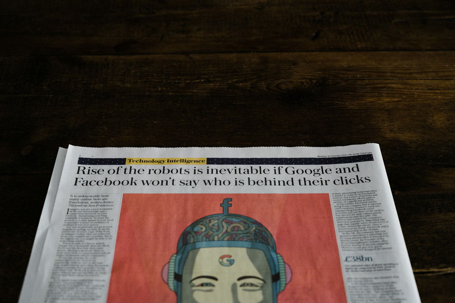 Newspaper with a headline about robots and tech companies on a wooden table. Features colorful illustration of a robot head.