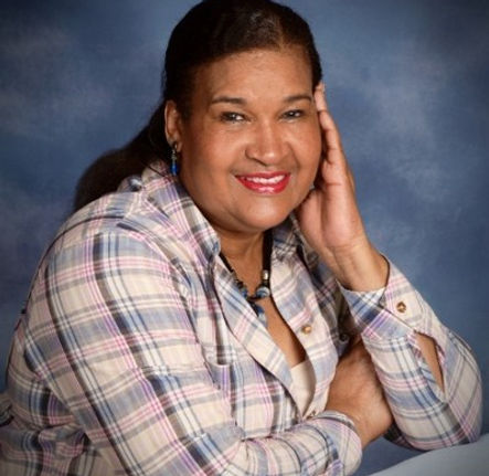 (Image Description: A photography portrait of Mashariki Sanyika from the waist up set against a cloudy, bluish gray photography background screen. Mashariki is wearing a multi-colored plaid long-sleeve button down shirt, a cream-colored undershirt, and chunky black jewelry. They are sitting with their arms crossed, one hand touching the side of their face. They are wearing light red lipstick and smiling at the camera.)