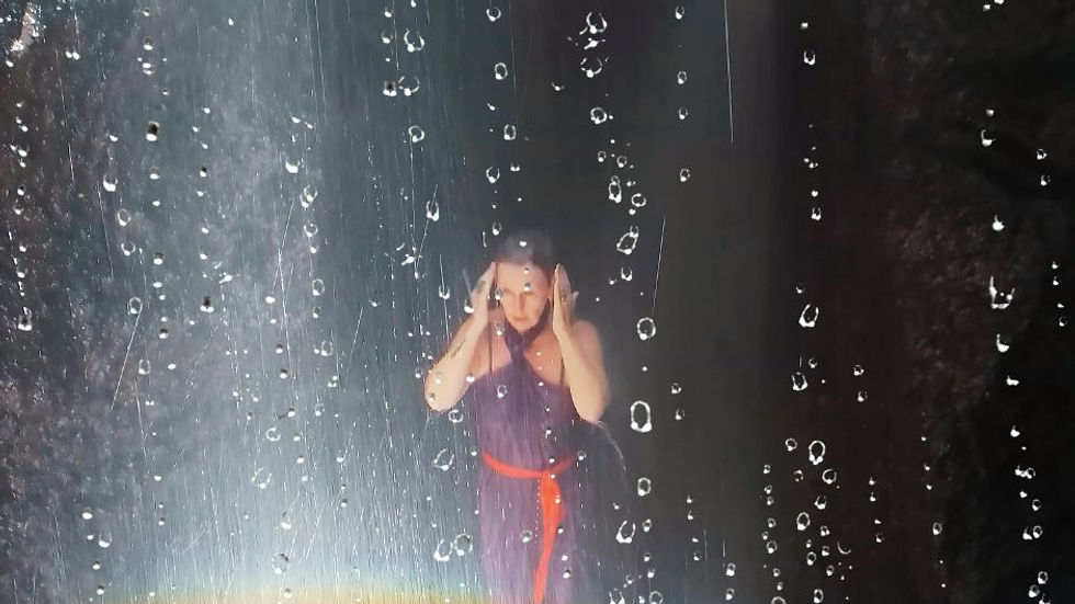 Person stands under waterfall, wearing a purple dress with red belt, hands on head. Water droplets and rainbow create a serene mood.