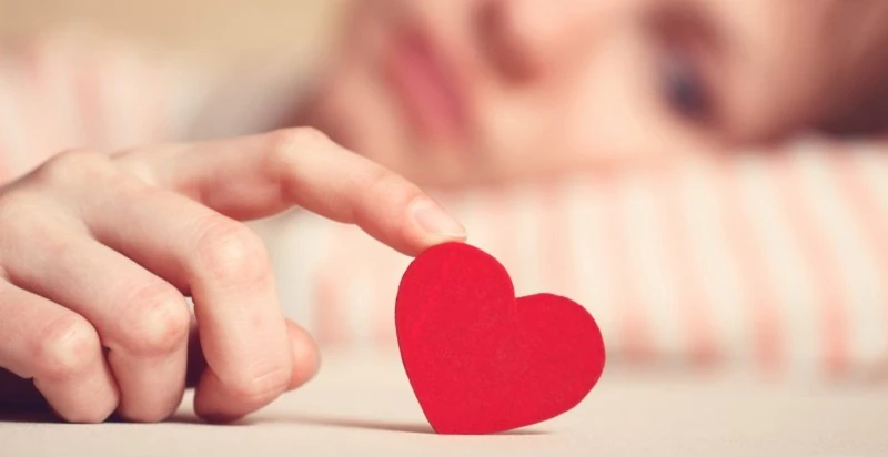 Person gently touching a small red heart while lying down, symbolizing emotional attachment.