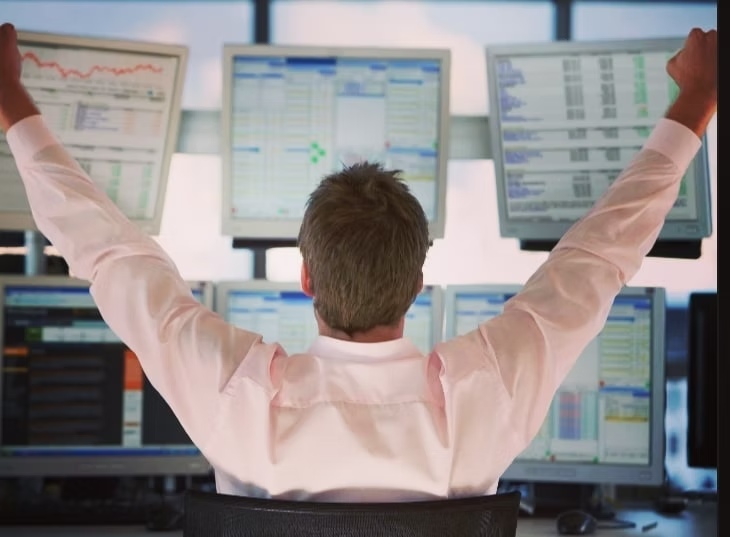 A man raises arms triumphantly at a desk with six monitors displaying financial charts in a bright office setting.