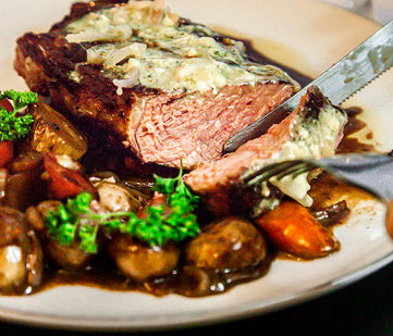Image of someone cutting into a juicy steak topped with blue cheese butter plated with marble potatoes, carrots, demi glaze.