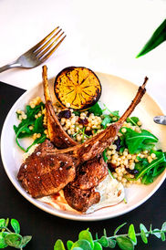 Image of a plate on a dark background with two lamb shanks, couscous, arugula, yogurt sauce, and a grilled lemon slice
