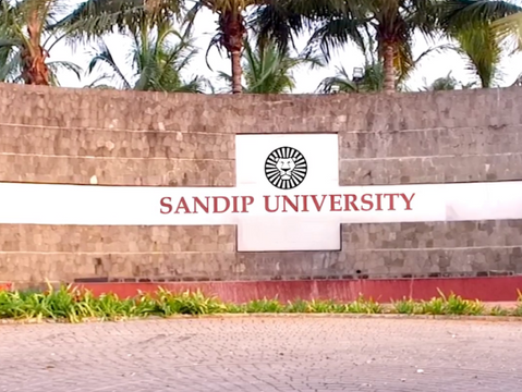 Gray brick wall with "SANDIP UNIVERSITY" text in red. Palm trees in the background and green shrubs in front. Circular emblem visible.
