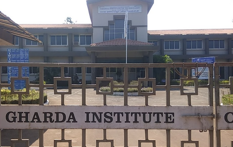Gate with "GHARDA INSTITUTE" text in front of a gray building with red roof. Clear sky and some greenery in the background.