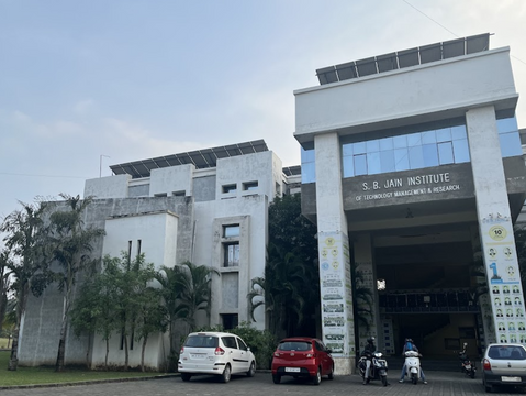 Modern white building of S.B. Jain Institute with blue-tinted windows. Three cars, two scooters parked in front, palm trees surround.