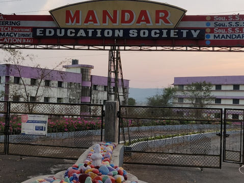 Entrance of Mandar Education Society. Sign with school names above metal gate. Purple-accented buildings in background, colorful stones below.