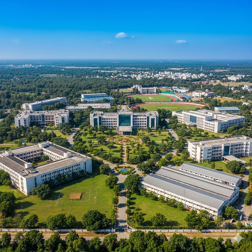 Aerial view of a vast campus with multiple modern buildings, lush green lawns, and a sports field. Clear blue sky in the background.