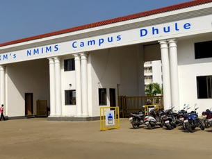 White building with columns, labeled SVKM's NMIMS Campus Dhule. Several motorcycles parked outside. Clear sky and one person walking.