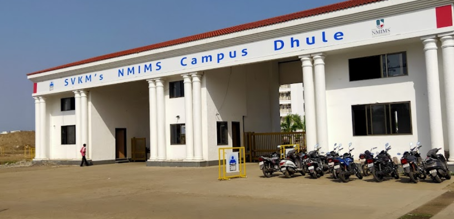 White building with columns, labeled SVKM's NMIMS Campus Dhule. Several motorcycles parked outside. Clear sky and one person walking.