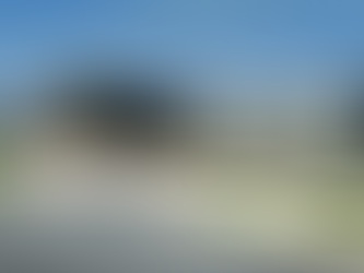 Modern building with large windows, surrounded by greenery. People walk toward the entrance on a paved path under a clear blue sky.