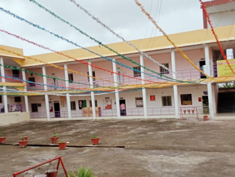 School courtyard with colorful streamers overhead. Yellow building, potted plants, and educational posters visible. Overcast sky.