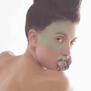 Creative shoot with white model with hair in a mohawk and green paint in her face posing with jewelry in her mouth