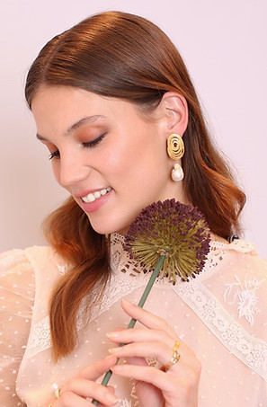 Bride with light brown hair with waves and soft makeup in brown tones wearing big pearls earrings and posing with flower