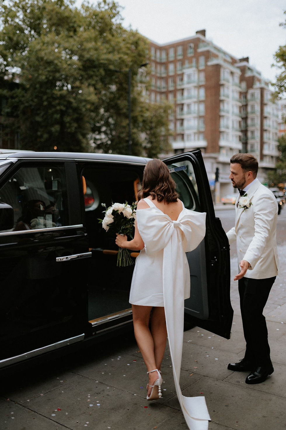 Bride in mini dress with big bow getting into London taxi with groom in white tux.