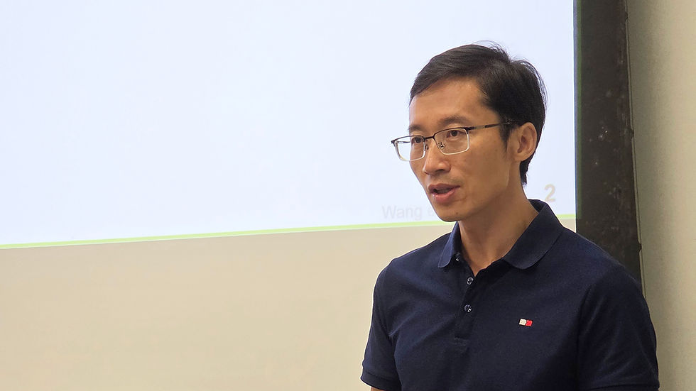 Man in navy polo and glasses speaking beside a projected slide with faint text Wang and 2 in a classroom or meeting room