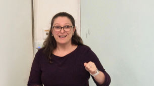 Smiling woman with glasses raises a fist indoors against a plain light wall and door, looking excited and cheerful