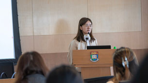 Woman speaking at a podium in a lecture hall, with a university crest and audience blurred in the foreground.