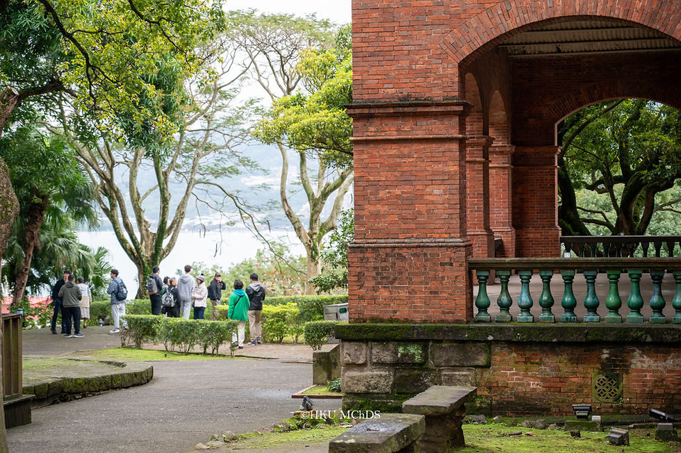 Tourists stand in a leafy park beside a red brick archway, with trees and a lake view beyond; watermark HKU MCHDS.