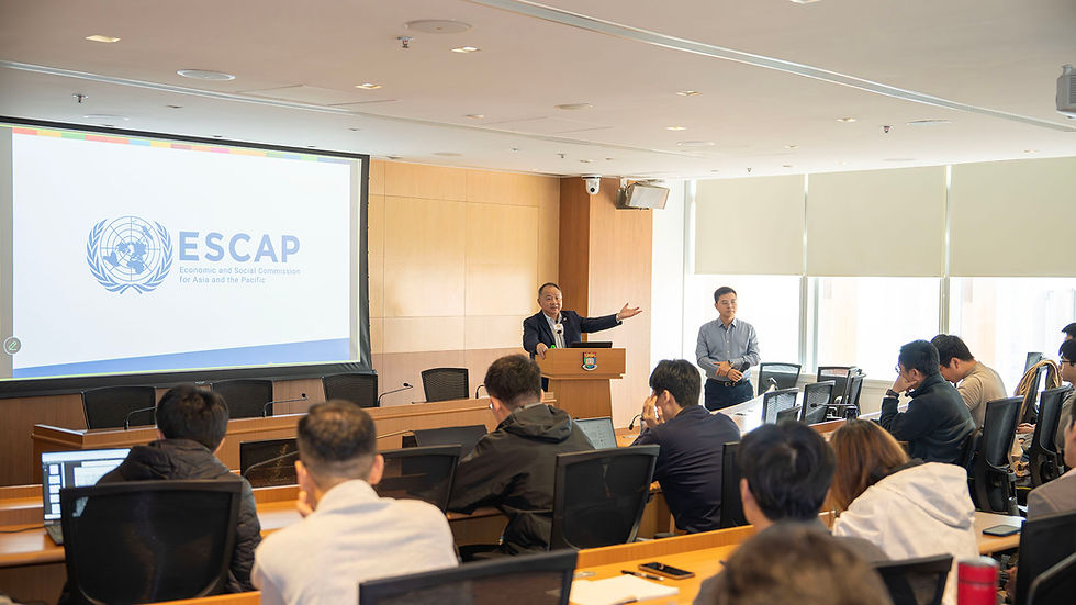 Speaker at podium addresses seated audience in lecture room; screen shows ESCAP logo and text, bright office setting.