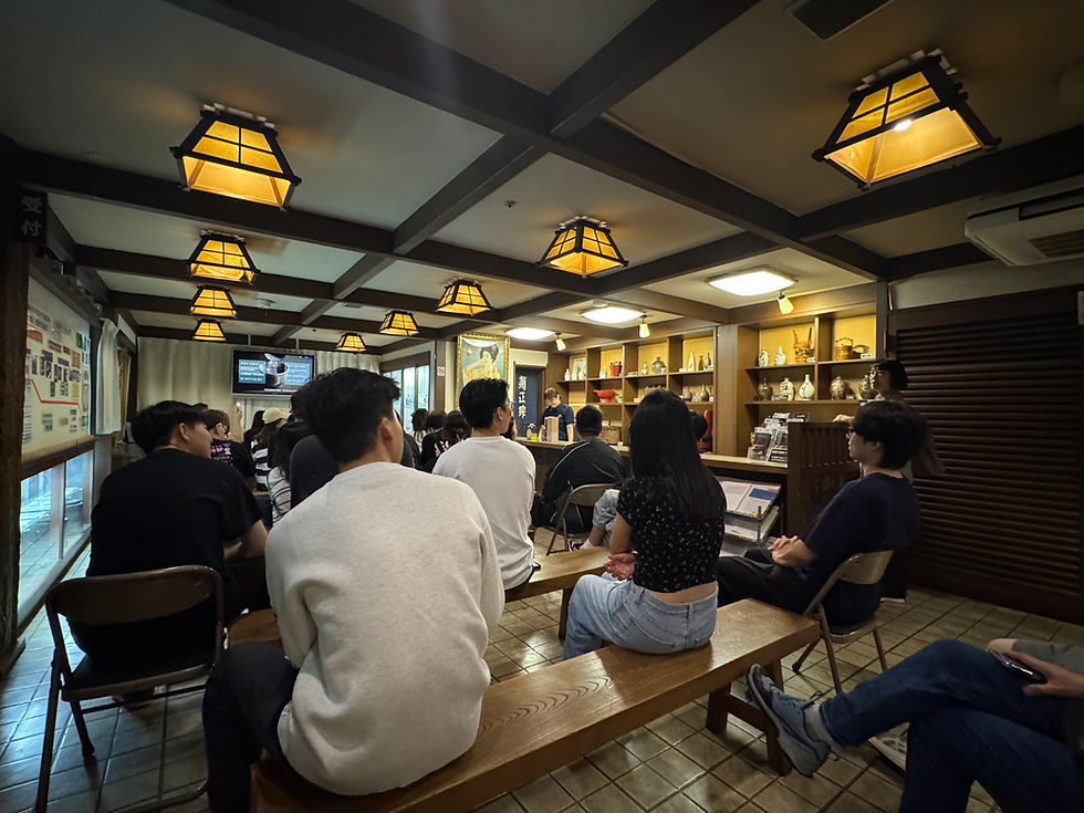 Crowded Japanese-style tea room with people seated on benches under warm lanterns, shelves of ceramics, and a screen with Japanese text.