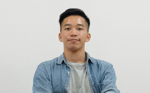 Young man in a denim shirt over a gray tee poses against a plain white wall, looking calm and neutral.