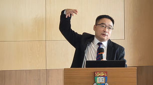 Man in a suit speaks at a podium, raising one hand beside a microphone and laptop, before a wood-paneled wall with a crest.