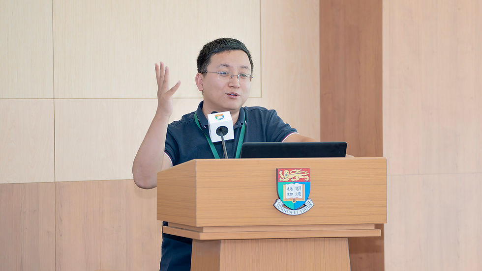 Man speaking at a wooden podium with a university crest, raising one hand beside a laptop and microphone in a wood-paneled room