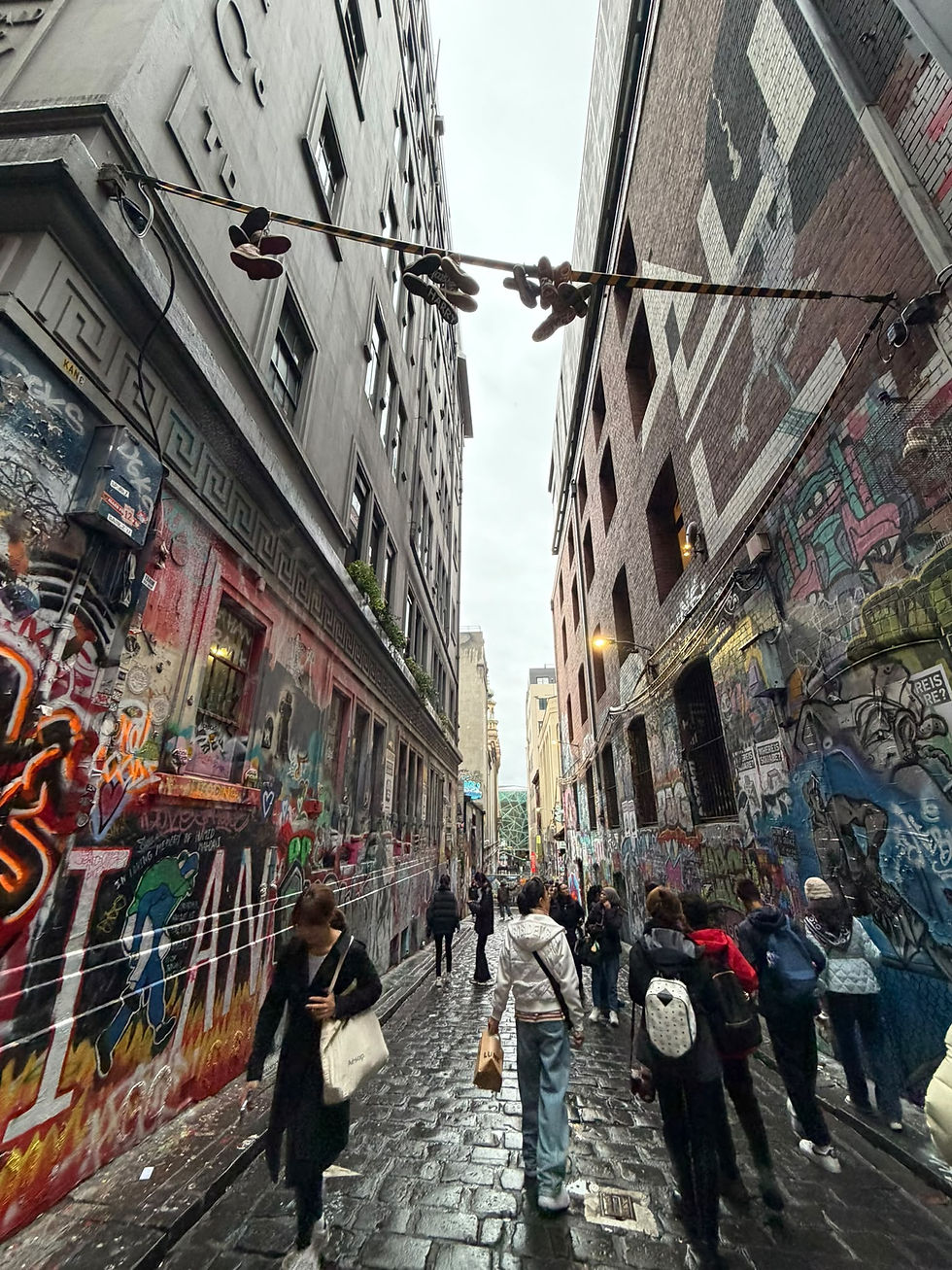People walk through a narrow wet graffiti-covered alley with hanging shoes overhead and tall buildings on both sides.