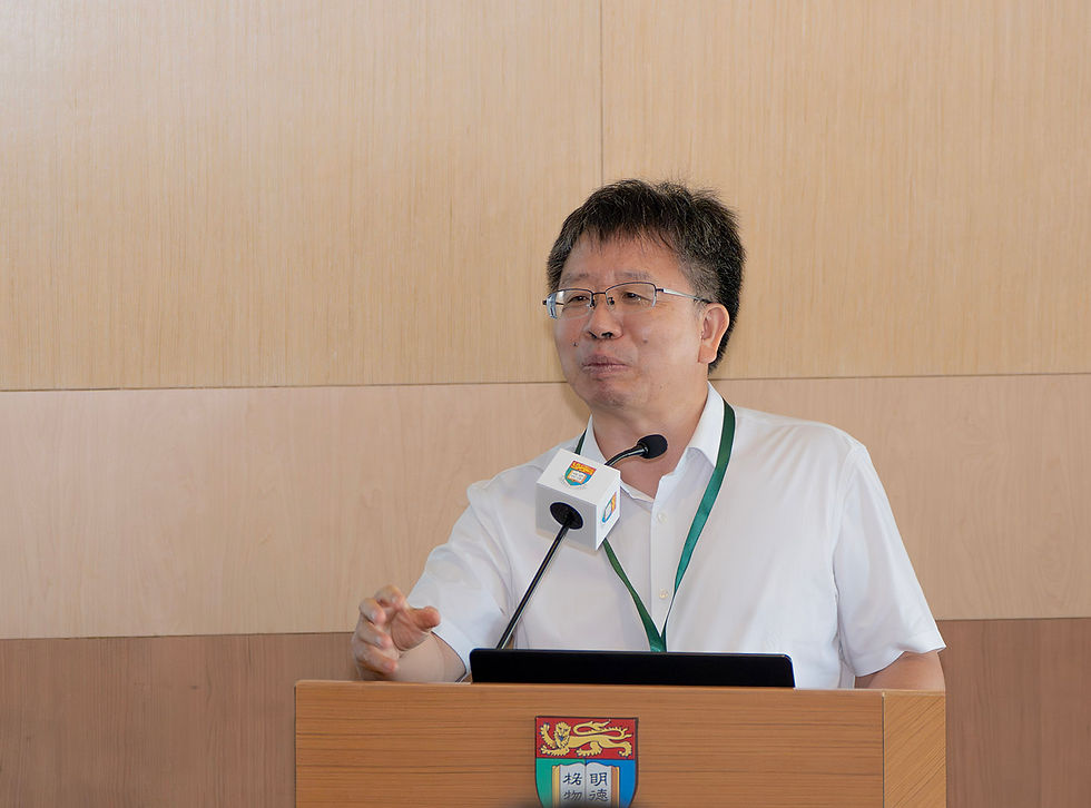 Man in glasses speaks at a podium with a microphone, wearing a white shirt and green lanyard against a wood-paneled wall.
