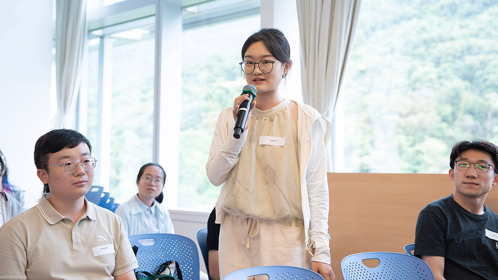 Young woman with glasses speaks into a microphone during a small meeting, seated listeners around her in a bright room.