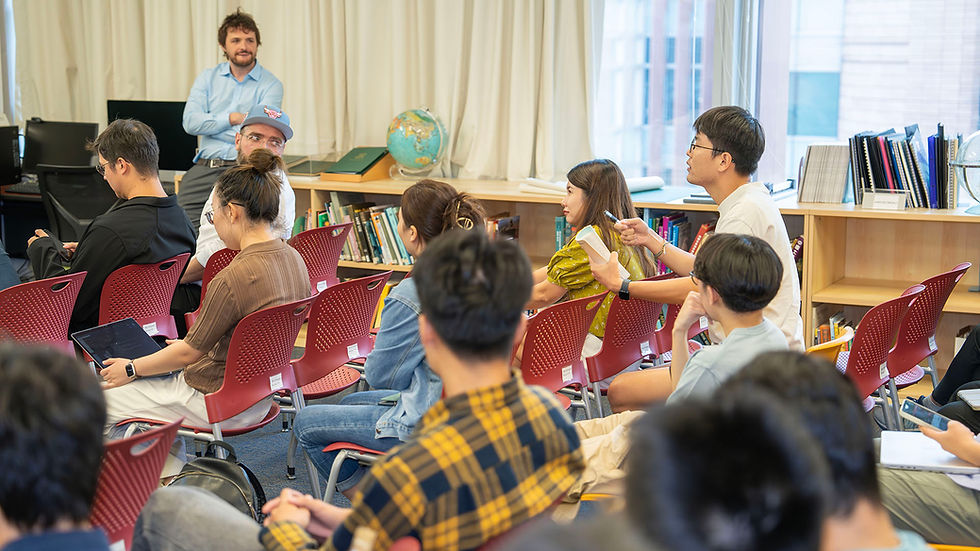 Students and a teacher discuss in a library classroom with red chairs, bookshelves, and a globe by bright windows.